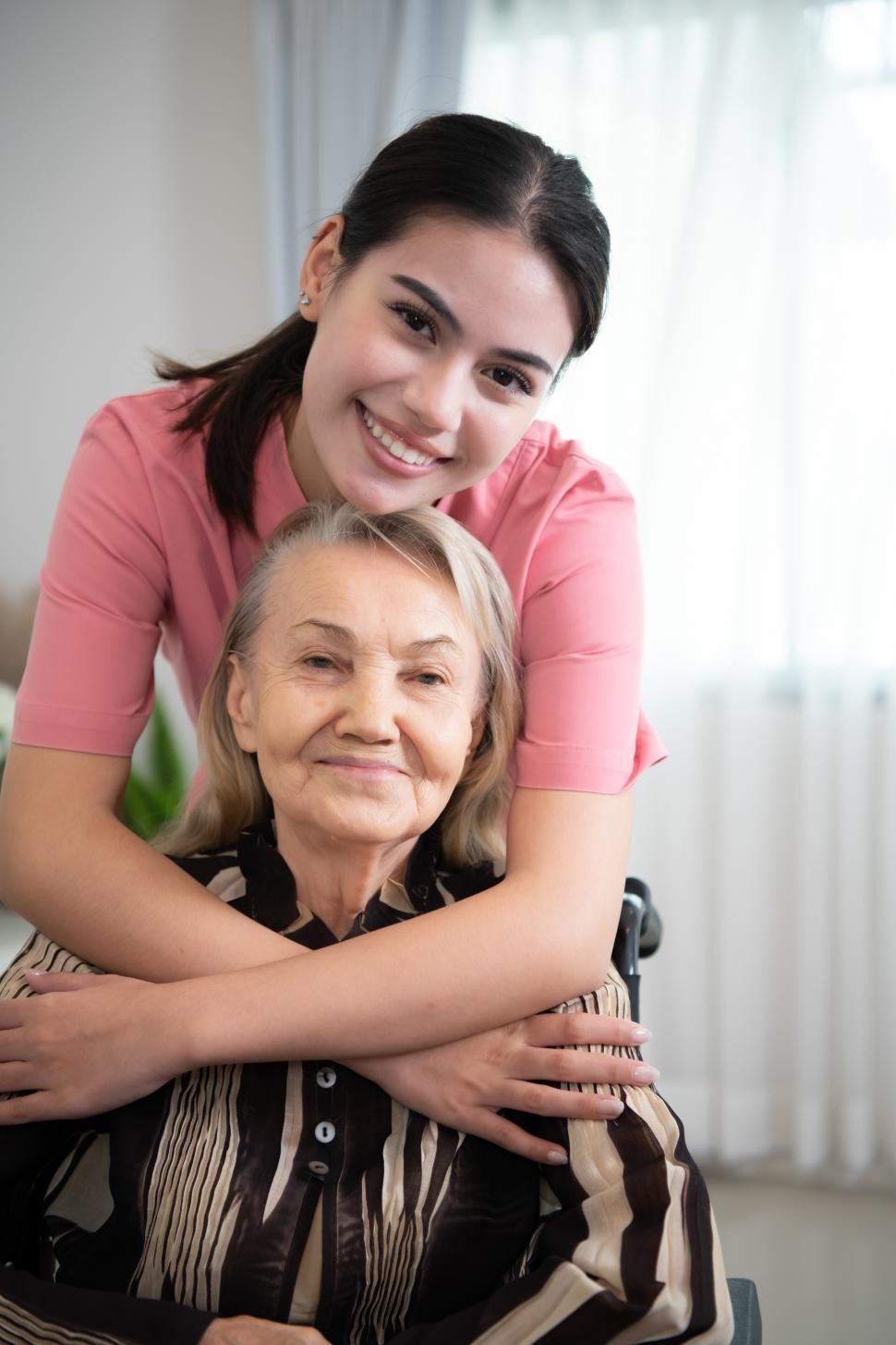 A young caregiver smiling and warmly embracing an older woman from behind