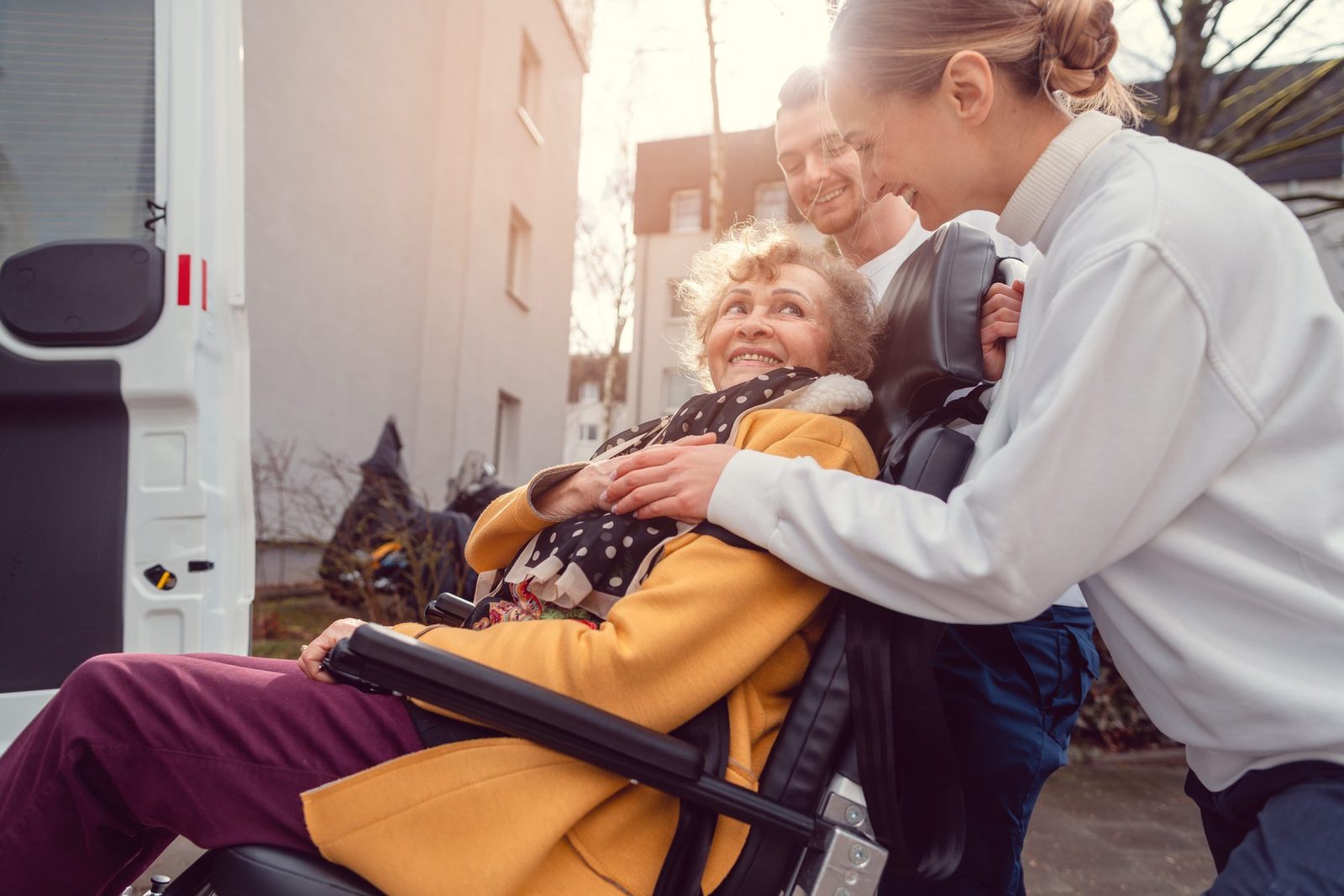 Two smiling caregivers helping a happy older woman in a wheelchair into an accessible van